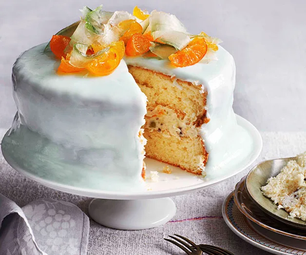 Cake with a light blue glaze, topped with candied fruit, a slice removed, placed on a white cake stand beside plates.