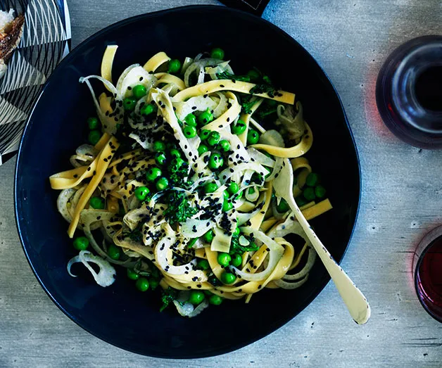 A black bowl with pasta, green peas, and herbs, garnished with black sesame seeds, placed on a gray table with red wine glasses.