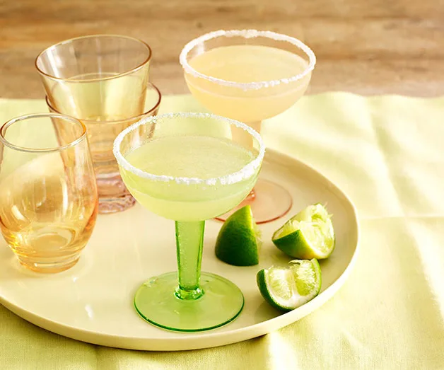 A tray with two margarita glasses with salt rims and lime wedges, alongside empty glasses on a yellow surface.