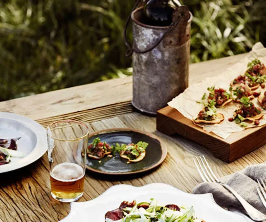 Rustic outdoor table with plates of food, a glass of beer, and a vintage metal jug, set against a grassy background.