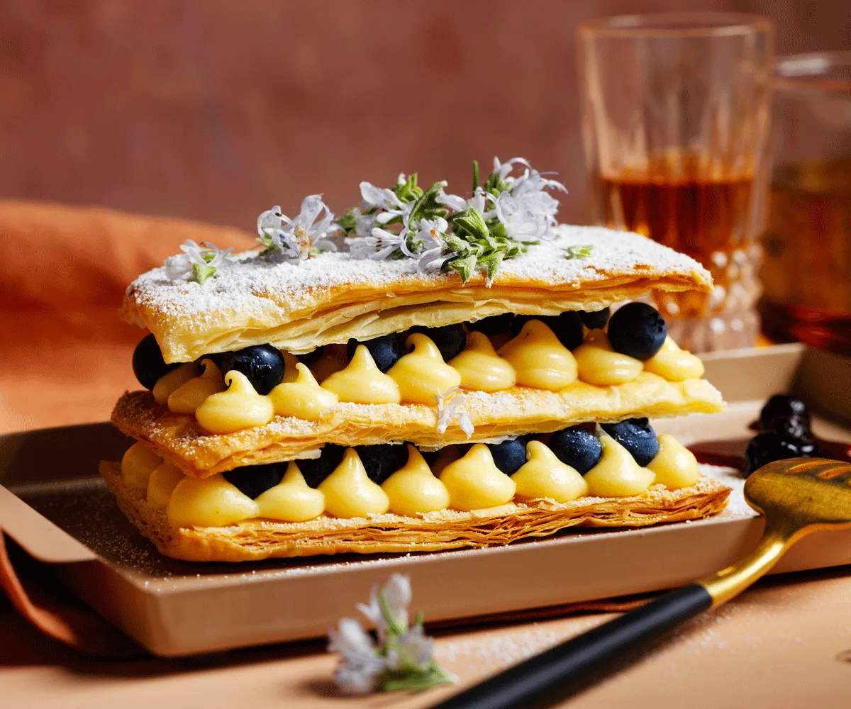 Milleufeuille with layers of crème pâtissière and blueberries and a glass of sweet vermouth in the background.