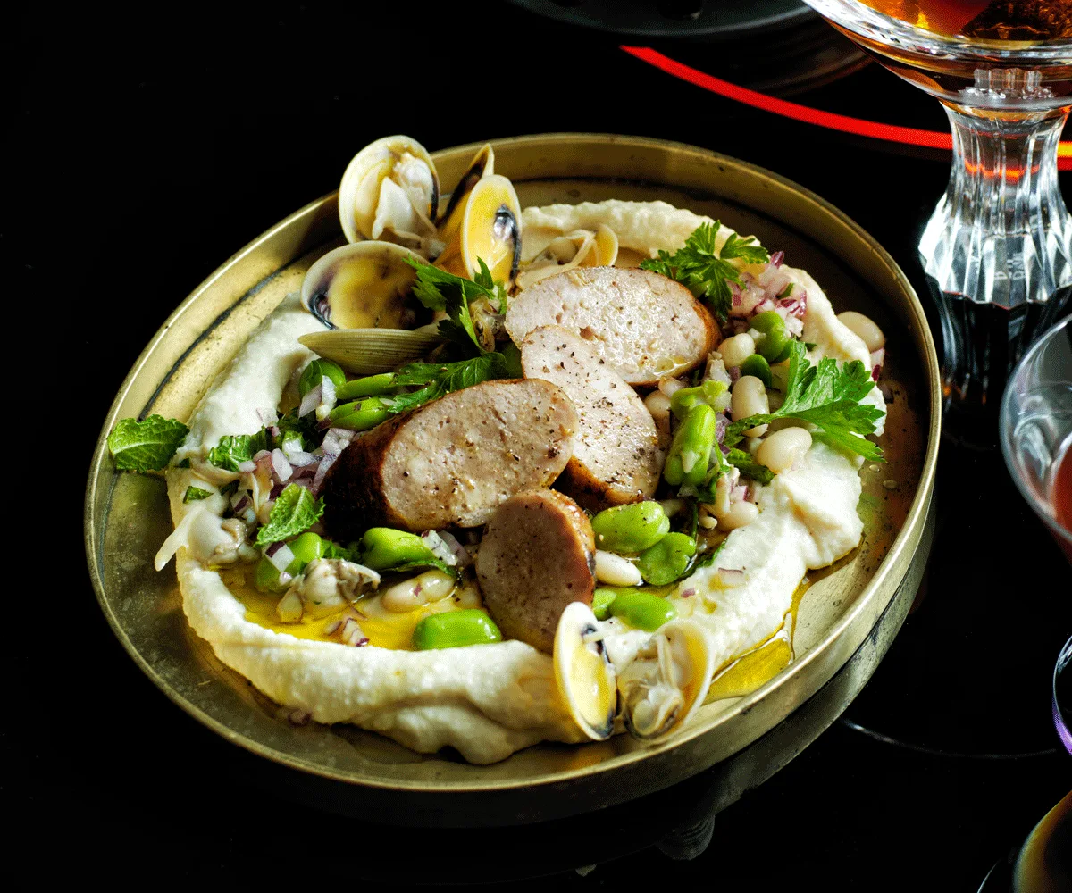 Botifarra sausage with clams and bean salad on a bed of white bean puree and served on a golden dish.