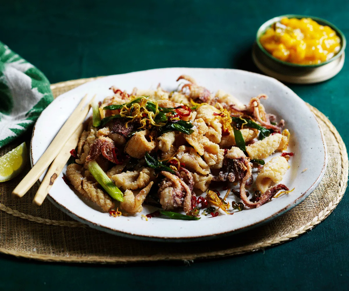 Spiced squid with spring onions on a white plate with chopsticks. In the background, a green dish filled with mango sweet and sour sauce.