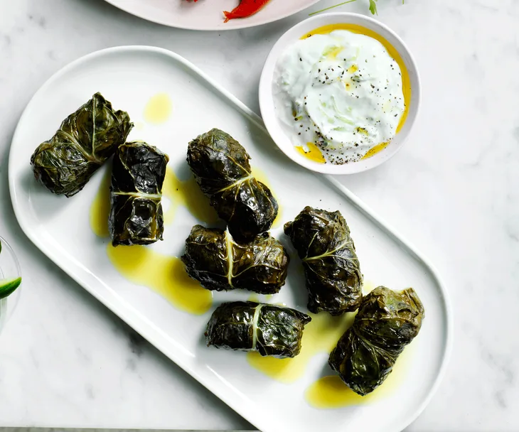 Dolmades wrapped in a silverbeet leaf served on a white platter and drixxled with olive oil. Tzatziki served on the side for dipping.