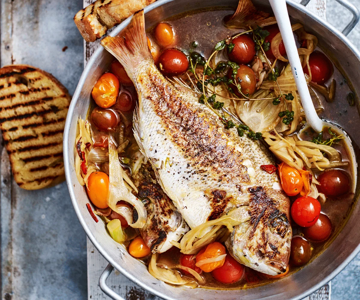 Whole baby snapper in a pan with tomato, fennel, and basil sauce. Large slices of charred sourdough on either side of the pot.