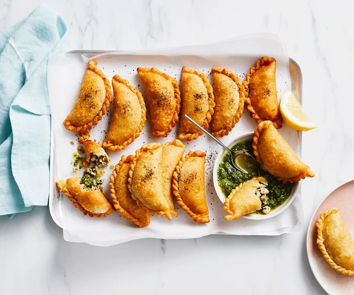 14 small empanadas on a white rectangular plate with mojo verde dipping sauce. Two are broken open with sauce drizzled over.