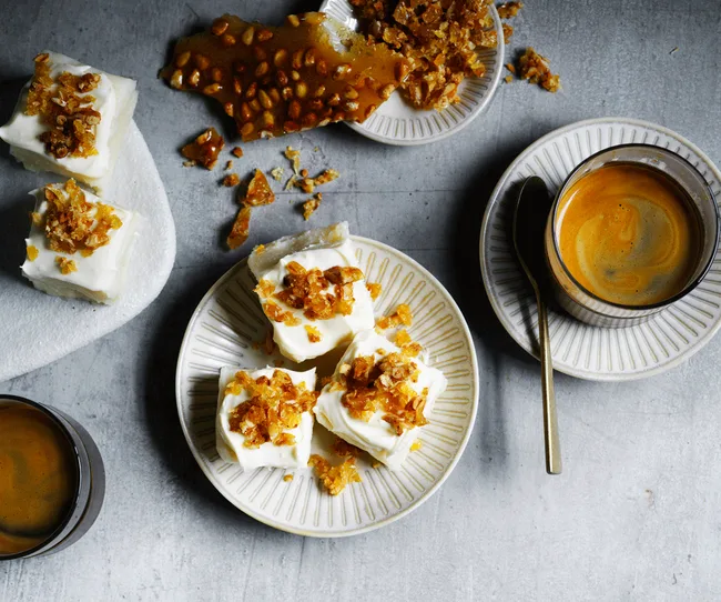 Squares of dessert topped with caramel on a plate, next to cups of coffee on a grey surface.