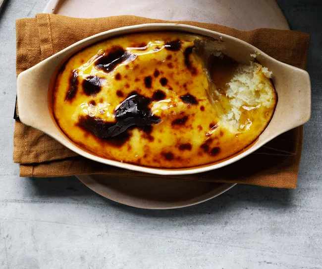 A ceramic dish with creamy baked rice pudding, topped with golden caramelized spots, placed on a brown cloth napkin.