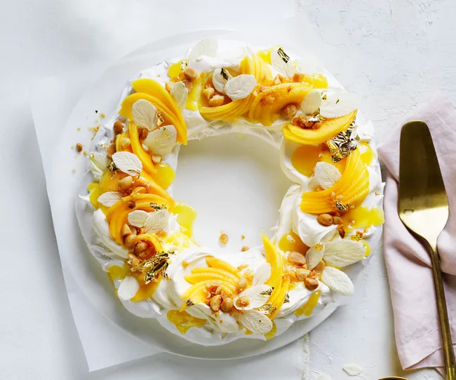 A round pavlova topped with mango slices, nuts, white chocolate, and gold flakes, on a white plate with a cake server.