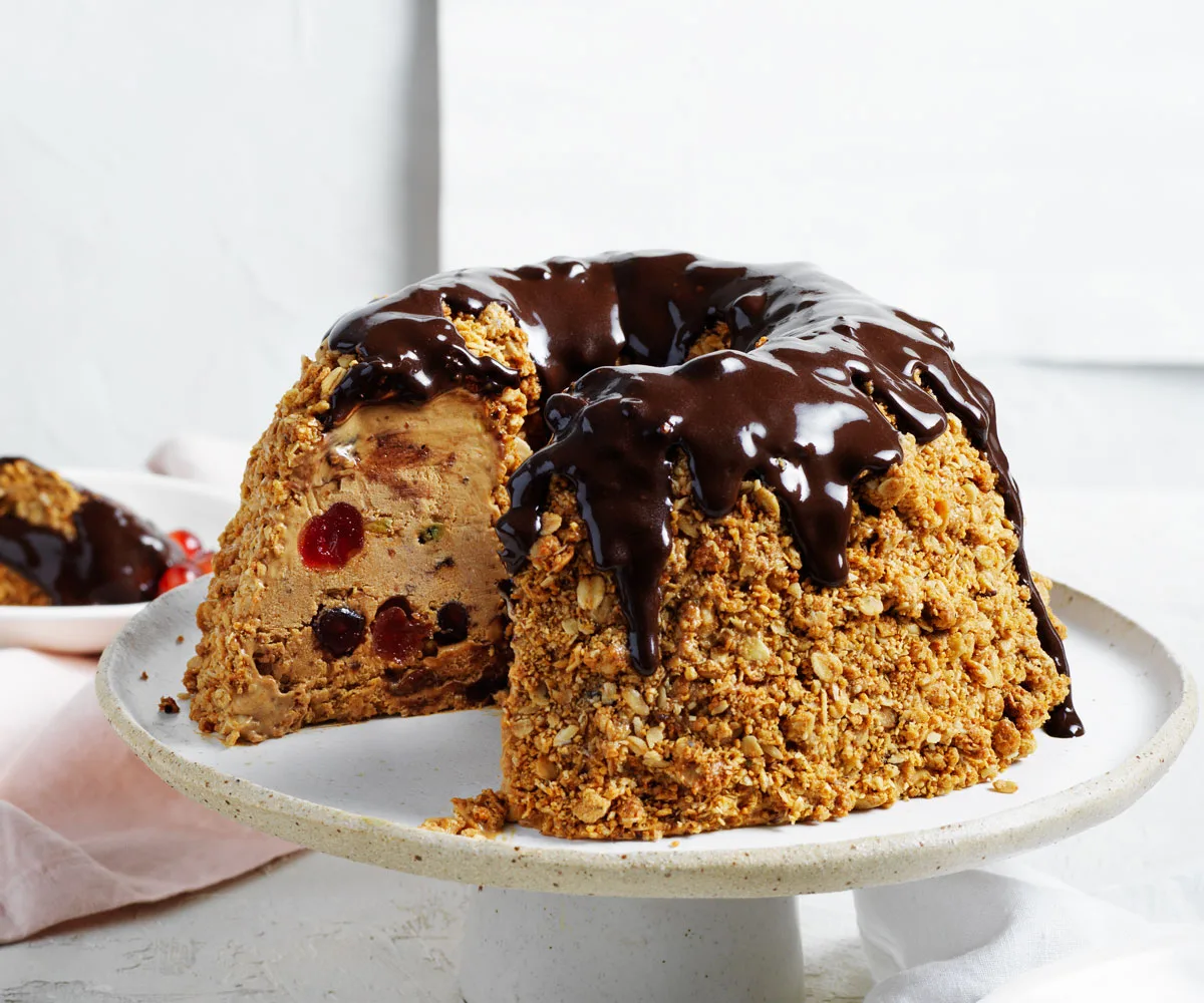 Photo of a white ceramic cake stand with crumbed Christmas ice-cream cake topped with glossy chocolate and whisky sauce