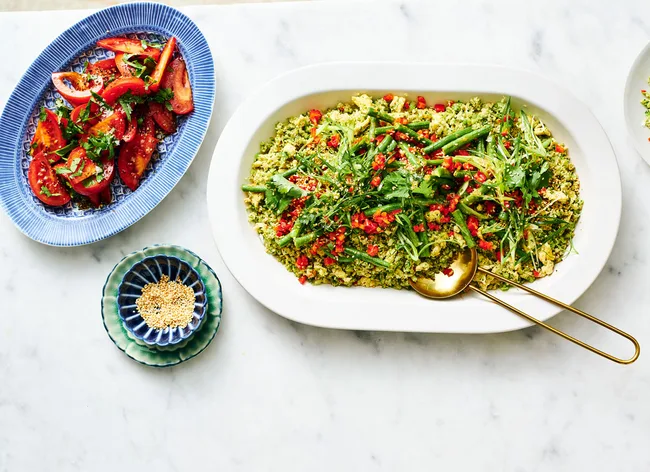 A bowl of quinoa salad with greens and red peppers, a side of sliced tomatoes, and a dish of sesame seeds on a white table.