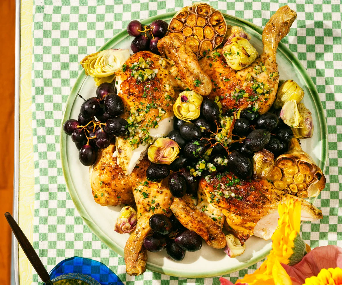 Bird's eye view of cooked chicken pieces with black grapes, roasted garlic halves and artichokes atop agreen checkered tablecloth