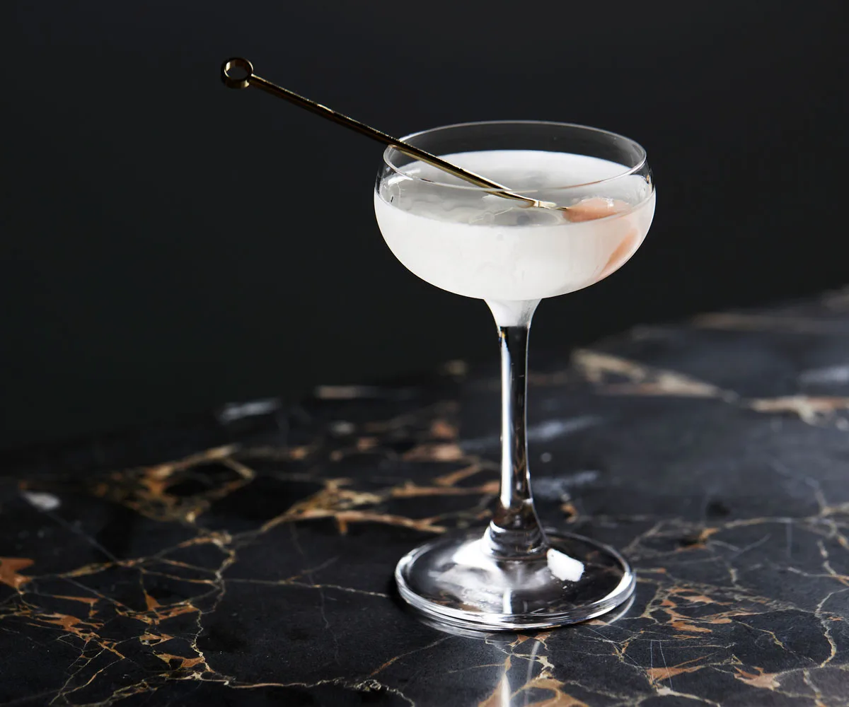 photo of black marble surface and a cocktail coupe with icy white liquid and metal skewer with pink garnish