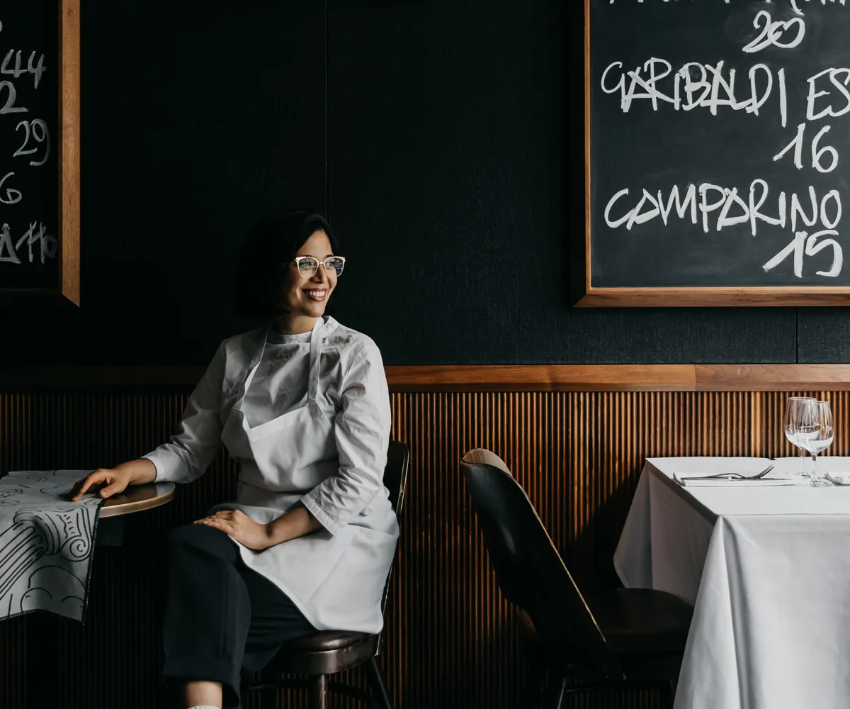 Portrait image of female chef in white apron sitting at restaurant table with blackboards with chalk writing in background