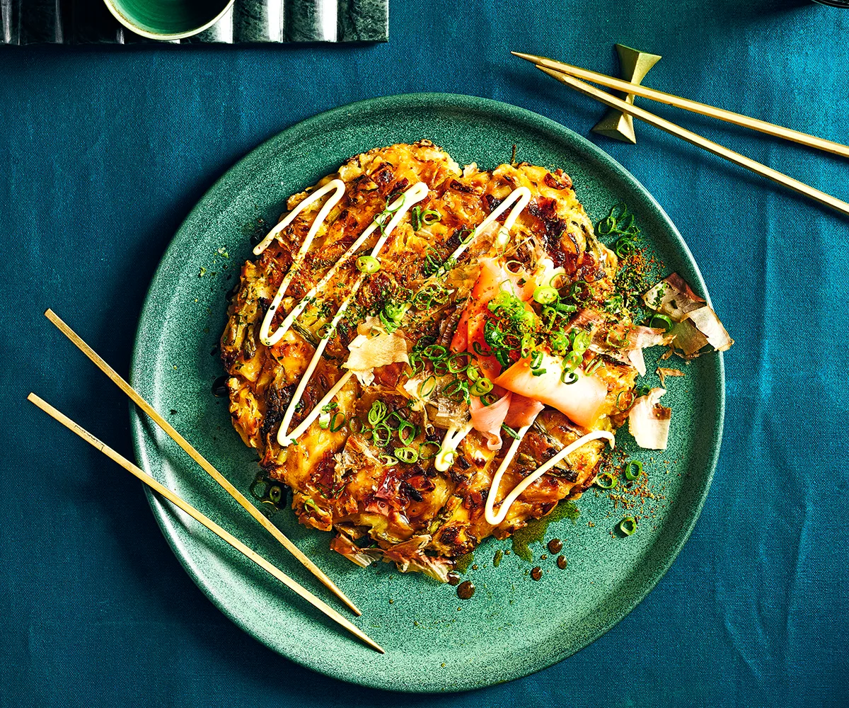 Overhead shot of a single okonomiyaki (Japanese pancake) on a green plate with chopsticks and blue cloth in frame