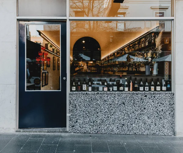 Street view of wine bar Leigh Street WIne Room, with a glass window, lined with wine bottles, looking into a long bar with a curved ceiling, and a navy door open ajar.