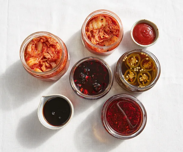Over the top shot of jars containing kimchi, teriyaki sauce, textural XO sauce, Korean hot sauce and pickled pickled jalapeños o a white surface. The jars cast long shadows on the surface.