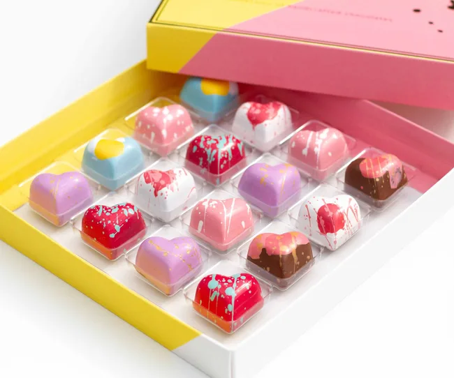 Box of colorful heart-shaped chocolates with artistic designs in an open yellow and pink box.