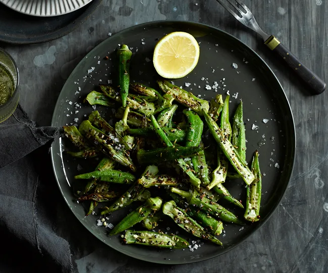 Grilled okra with lemon and spices on a dark plate, garnished with sea salt, with a fork nearby.