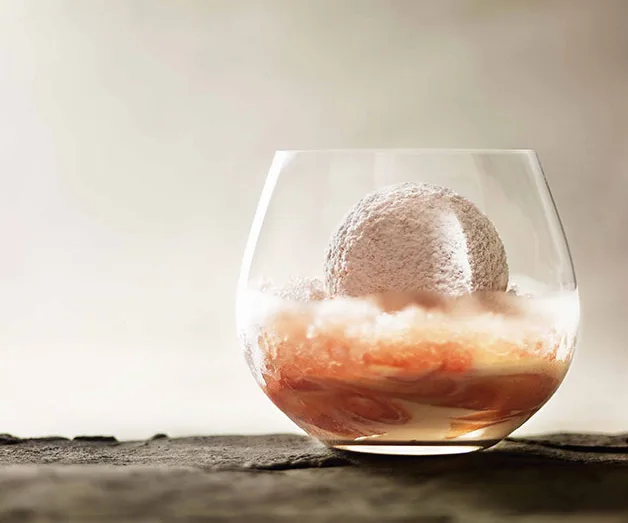 Clear glass with a white dessert sphere on a bed of pink jelly, against a light brown background.
