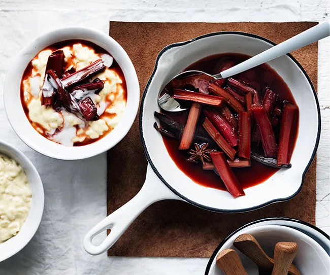 White bowl of vanilla rice pudding with roasted rhubarb next to a pan of rhubarb in red syrup.