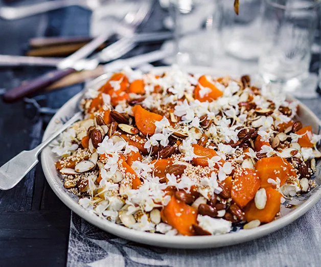 Plate of salad with sliced persimmons, nuts, seeds, and white flower petals on a table setting.