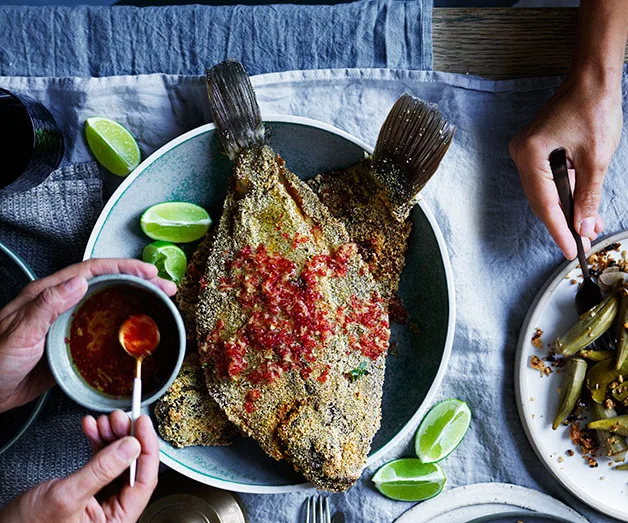 Crispy fried fish topped with chili, surrounded by lime wedges, with hands holding sauce and eating from a plate.