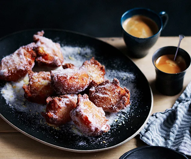 Golden sugar-dusted apple fritters on a black plate, served with two cups of coffee on a wooden table.
