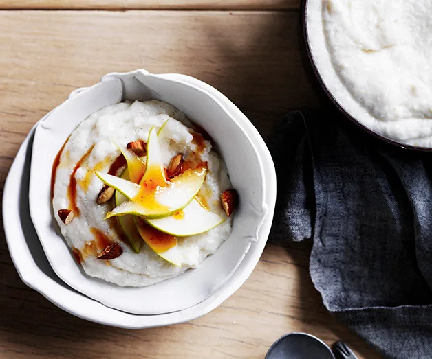 Creamy porridge topped with sliced pears, almond pieces, and syrup in a white bowl on a wooden table.