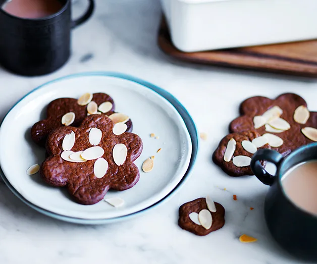 Flower-shaped chocolate cookies with almond slices on a plate next to a mug of hot beverage.