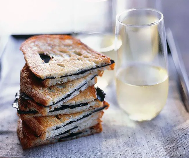 Stack of toasted truffle sandwiches with glasses of white wine on a table.