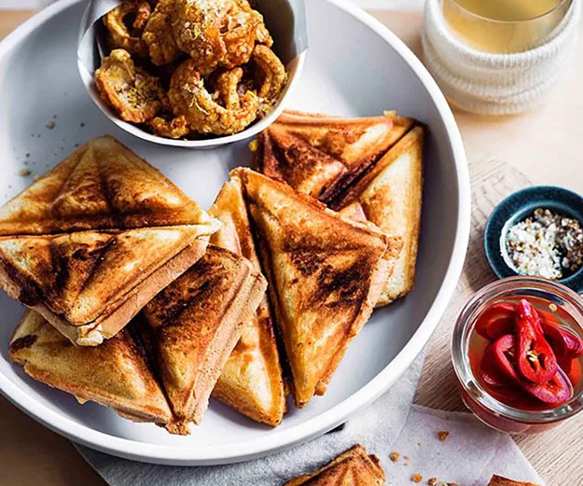 Triangular toasted sandwiches with a bowl of pork cracklings, sliced pickles, and a drink on a table.