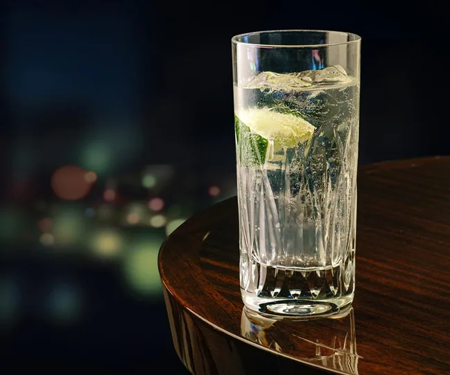A glass of gin and tonic with ice and lime wedge on a wooden table, with a blurred bokeh background.