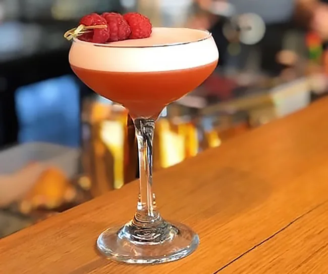 A cocktail in a coupe glass with a frothy top, garnished with raspberries, on a wooden bar counter.