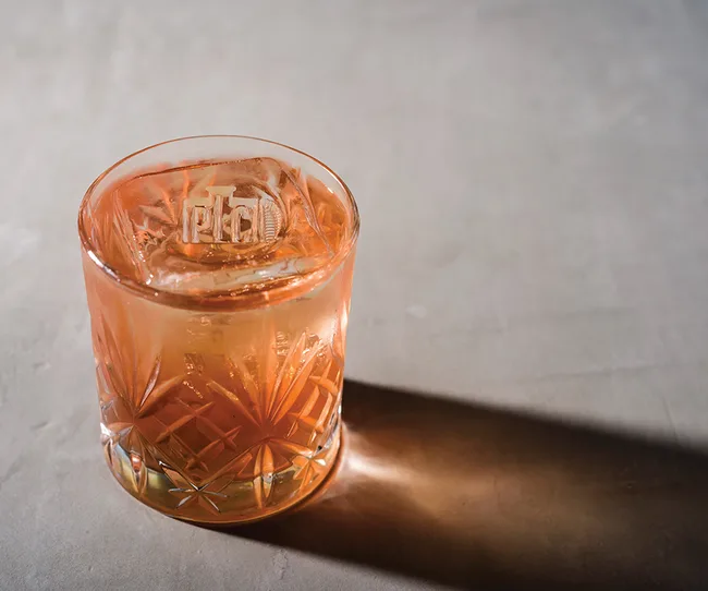 A glass of peach-colored cocktail with ice on a textured surface, casting a shadow.