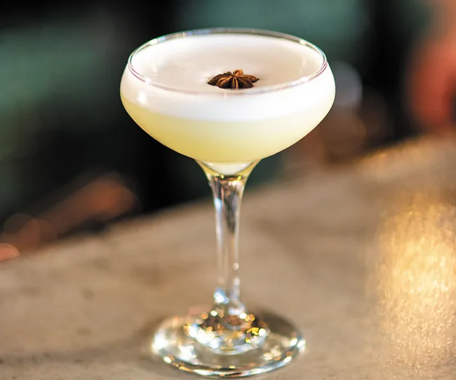 A cocktail with a foamy top garnished with star anise in a coupe glass on a bar counter.
