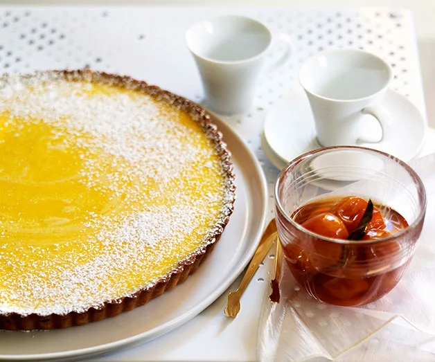 Lemon tart dusted with powdered sugar, served with poached tamarillos and two white cups on a white table.