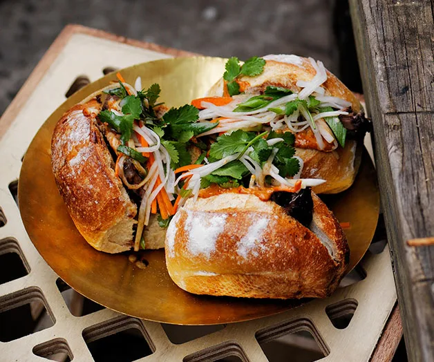 Three banh mi sandwiches on a brass plate, filled with pickled vegetables and cilantro.