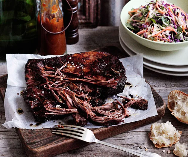 Slow-cooked beef brisket on a wooden board with a fork, accompanied by a bowl of coleslaw, sauce, and bread pieces.