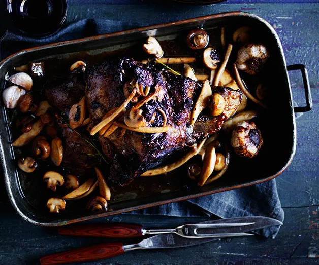 Roasted lamb with mushrooms and herbs in a baking pan, accompanied by a fork and knife on a table setting.