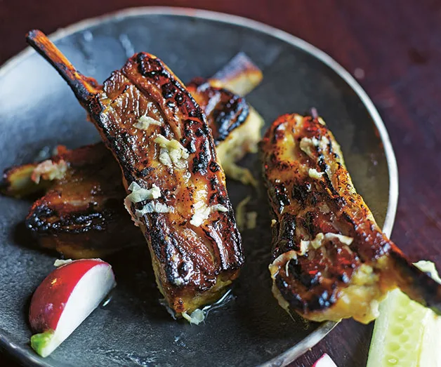 Grilled lamb ribs on a dark plate, garnished with garlic, served with a radish wedge and pickle slice on the side.