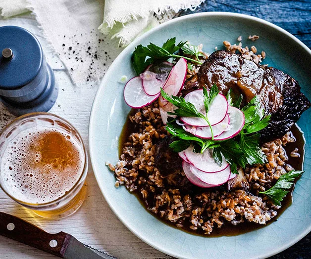 Braised beef cheeks on farro with radish and parsley, served with a glass of beer and a pepper mill on the side.