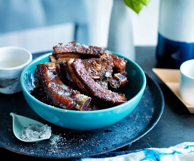 A bowl of glazed, seasoned pork ribs garnished with seasoning, placed on a round tray with cups nearby.