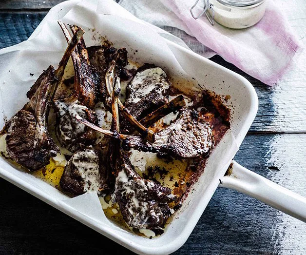 Grilled lamb cutlets in a white baking dish drizzled with white sauce, set on a rustic wooden table.