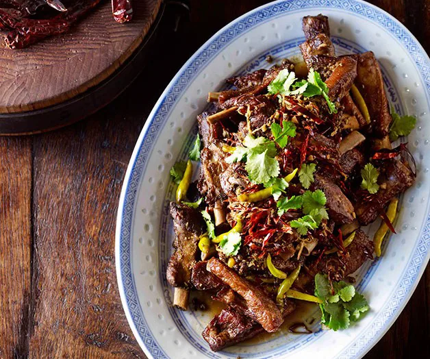 Plate of braised lamb ribs, garnished with cilantro and chilies, on a wooden table.