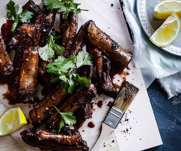 Barbecue beef ribs garnished with cilantro, served with lemon wedges on a wooden board.