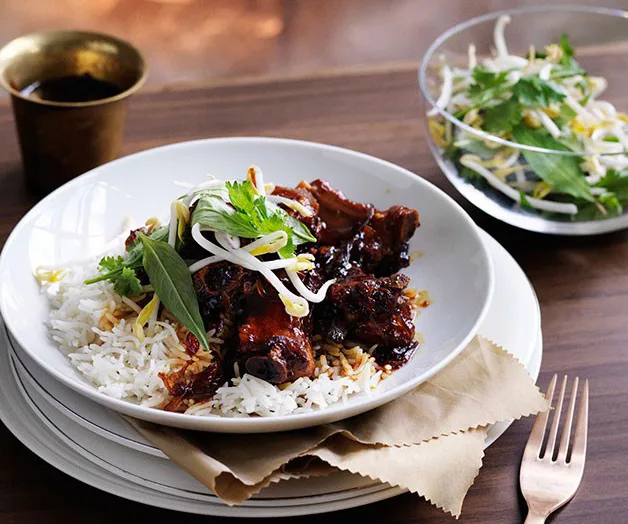 Braised pork ribs on rice with herbs and sprouts, accompanied by a side salad, on a wooden table with utensils.