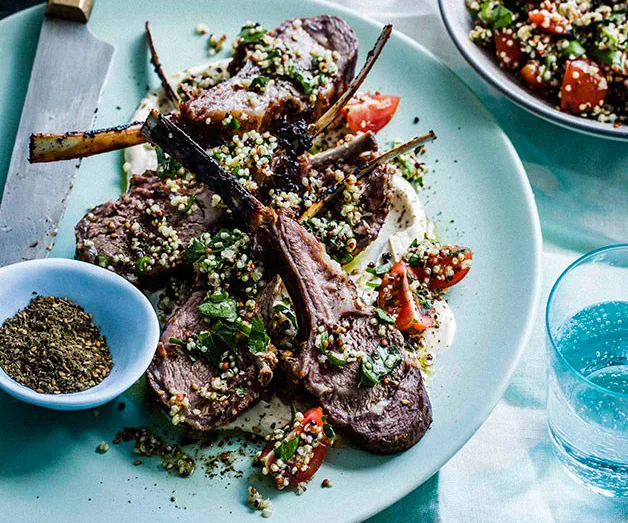Grilled lamb chops garnished with herbs and quinoa salad on a blue plate, with a side of spice and a glass of water.