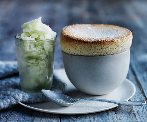 A lemon soufflé with a browned top in a gray cup on a white plate, served with a glass of lemon sorbet.