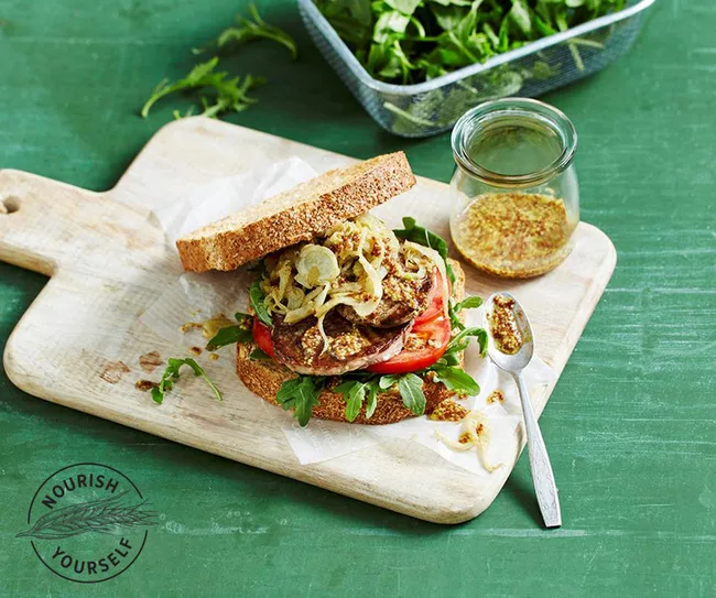 Open sandwich with grilled patty, tomato, arugula, and onions on a wooden board, with mustard in a jar.